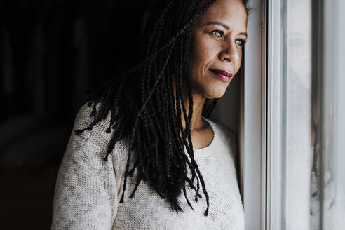 Contemplating woman looking through window while standing at home - EBBF00958