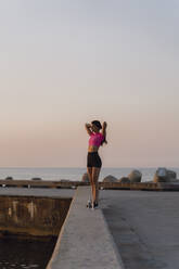 Junge Frau mit Hand in den Haaren steht auf einer Stützmauer am Meer - AFVF07354