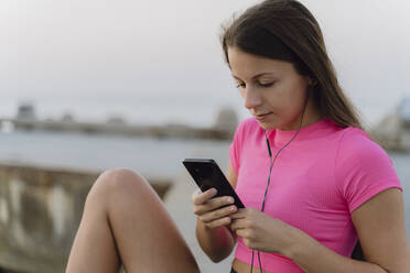 Sportler mit In-Ear-Kopfhörern, der ein Mobiltelefon benutzt, während er auf einer Stützmauer am Meer sitzt - AFVF07358