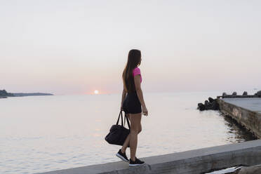 Sportlerin hält Tasche, während sie auf der Stützmauer gegen das Meer läuft - AFVF07360