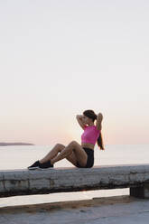 Athlete tying hair while sitting on concrete wall against sea - AFVF07366