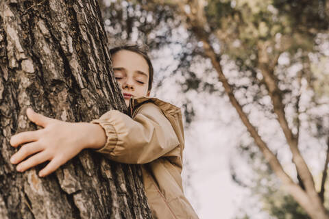 Nettes Mädchen mit geschlossenen Augen umarmt Baum im Park, lizenzfreies Stockfoto