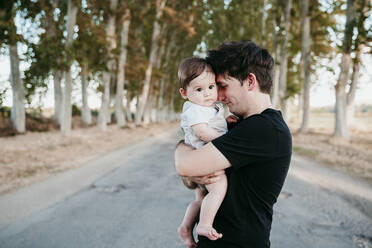 Smiling father embracing cute baby while standing on road outdoors - EBBF00981