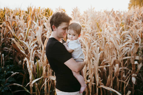 Vater trägt kleinen Jungen, während er bei Sonnenuntergang vor einem Feld steht - EBBF00984