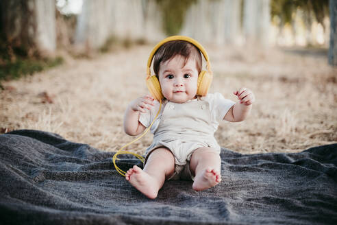 Niedlicher kleiner Junge trägt Kopfhörer, während er auf einer Decke im Freien sitzt - EBBF00986