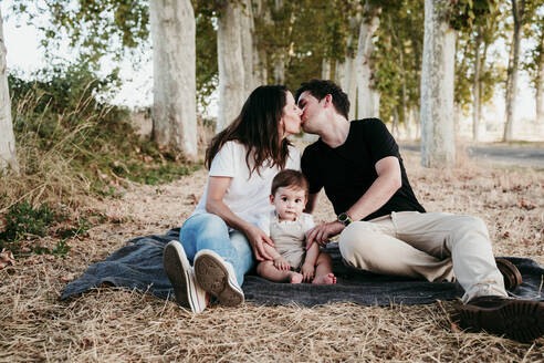 Eltern, die sich küssen, während sie mit Baby Bot auf einer Decke im Freien sitzen - EBBF00992