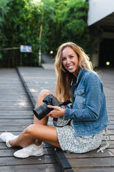 Lächelnde schöne blonde Frau, die eine Digitalkamera hält, während sie auf einem Fußweg sitzt - JMPF00474