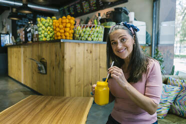 Smiling mature woman holding fresh juice in mason jar while sitting at table - XLGF00622