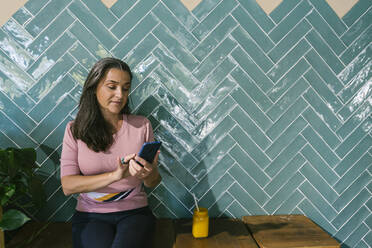 Woman using smart phone while sitting with juice against turquoise wall at bar - XLGF00640