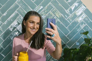 Lächelnde Frau, die ein Selfie macht, während sie einen Saft gegen die türkisfarbene Wand eines Cafés hält - XLGF00641