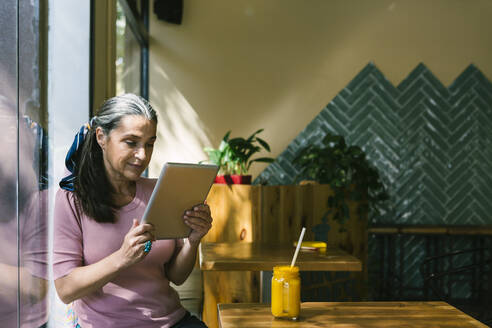 Frau hält digitales Tablet und lehnt sich an das Fenster eines Cafés - XLGF00646