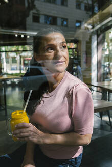 Nachdenkliche Frau, die ein Einmachglas mit Saft hält, während sie durch das Fenster eines Cafés schaut - XLGF00648