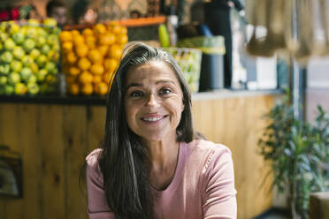 Smiling mature woman sitting against fruits at bar - XLGF00655