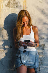 Lächelnde Frau, die ein Mobiltelefon benutzt, während sie an der Wand steht - JMPF00479