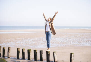Happy young woman standing on wooden post with arms raised at beach against clear sky during sunny day - UUF21747