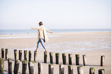 Junge Frau mit ausgestreckten Armen, die an einem sonnigen Tag am Strand auf Holzpfählen läuft - UUF21748