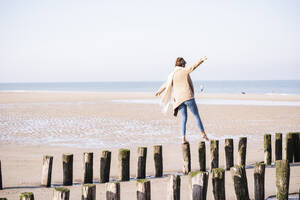 Junge Frau, die es genießt, an einem sonnigen Tag am Strand auf Holzpfählen mit ausgestreckten Armen zu laufen - UUF21749