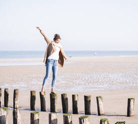 Lächelnde junge Frau mit ausgestreckten Armen, die an einem sonnigen Tag auf Holzpfählen am Strand spazieren geht - UUF21750