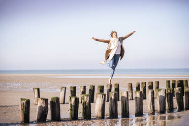 Fröhliche junge Frau mit ausgestreckten Armen, die an einem sonnigen Tag am Strand auf Holzpfählen läuft - UUF21751
