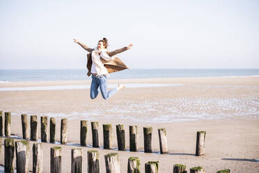 Happy young woman with arms outstretched jumping from wooden posts at beach during sunny day - UUF21753