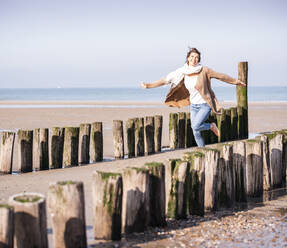 Smiling young woman with arms outstretched running amidst wooden posts at beach during sunny day - UUF21754