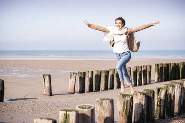 Cheerful young woman with arms outstretched running amidst wooden posts at beach during sunny day - UUF21755