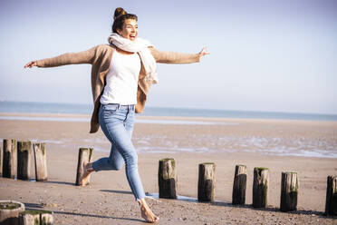 Happy young woman with arms outstretched running amidst wooden posts at beach during sunny day - UUF21756