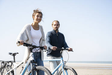 Young couple walking with bicycles at beach against clear sky on sunny day - UUF21761