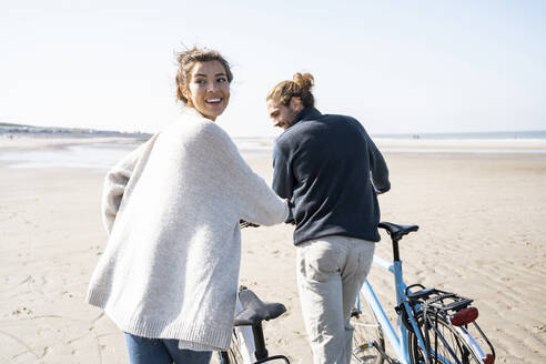 Lächelnde junge Frau, die mit Fahrrad und Freund spazieren geht, während sie über die Schulter auf den Strand gegen den klaren Himmel an einem sonnigen Tag schaut - UUF21762