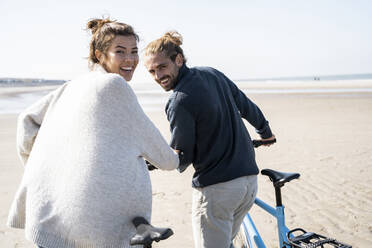 Glückliches junges Paar, das mit Fahrrädern am Strand gegen den klaren Himmel an einem sonnigen Tag spazieren geht - UUF21763