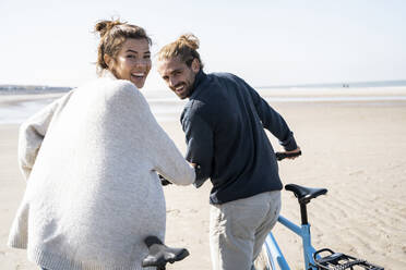 Happy young couple walking with bicycles at beach against clear sky on sunny day - UUF21763