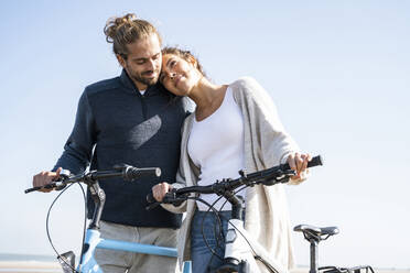 Beautiful woman with head on boyfriend's shoulder standing with bicycles at beach against clear sky on sunny day - UUF21764