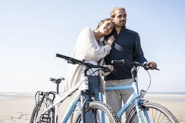 Smiling beautiful woman with head on boyfriend's shoulder standing with bicycles at beach against clear sky on sunny day - UUF21765