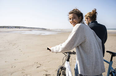 Smiling beautiful woman walking with bicycle by boyfriend while looking over shoulder at beach against clear sky on sunny day - UUF21766