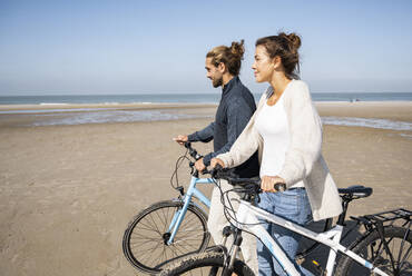 Young boyfriend and girlfriend walking with bicycles at beach against clear sky on sunny day - UUF21767
