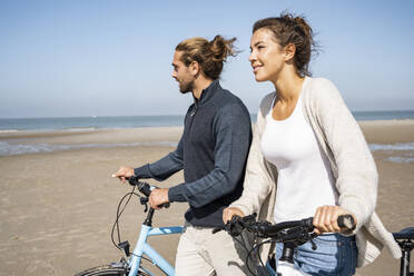 Smiling young woman walking with boyfriend and bicycles at beach against clear sky on sunny day - UUF21768