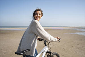 Cheerful young woman walking with bicycle while looking over shoulder at beach on sunny day - UUF21769