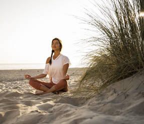 Beautiful young woman practicing yoga while sitting by plant on sand at beach against clear sky during sunset - UUF21770