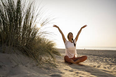 Beautiful young woman practicing yoga while sitting cross-legged by plant on sand at beach against clear sky during sunset - UUF21772