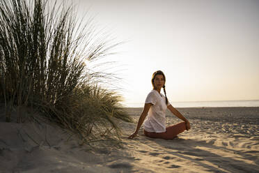 Beautiful young woman practicing yoga while sitting by plant at beach against clear sky during sunset - UUF21773