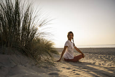 Schöne junge Frau übt Yoga, während sie am Strand gegen den klaren Himmel bei Sonnenuntergang sitzt - UUF21773