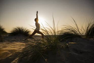 Silhouette young woman practicing warrior position yoga amidst plants at beach against clear sky during sunset - UUF21777