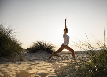 Junge Frau übt Yoga in der Kriegerposition inmitten von Pflanzen am Strand gegen den klaren Himmel bei Sonnenuntergang - UUF21778