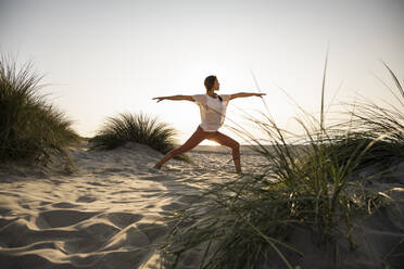 Junge Frau übt Krieger 2 Position Yoga inmitten von Pflanzen am Strand gegen klaren Himmel bei Sonnenuntergang - UUF21779