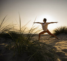 Junge Frau übt Krieger 2 Position Yoga am Strand gegen klaren Himmel bei Sonnenuntergang - UUF21780