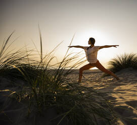 Young woman practicing warrior 2 position yoga at beach against clear sky during sunset - UUF21780