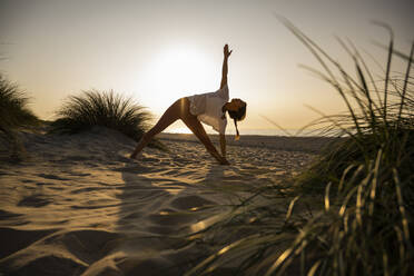 Junge Frau übt Yoga in Dreiecksposition inmitten von Pflanzen am Strand gegen den klaren Himmel bei Sonnenuntergang - UUF21781