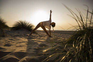 Young woman practicing triangle position yoga amidst plants at beach against clear sky during sunset - UUF21781