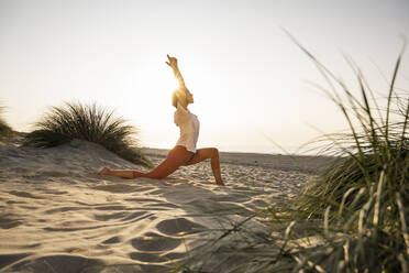 Young woman practicing yoga on sand at beach during sunset - UUF21782