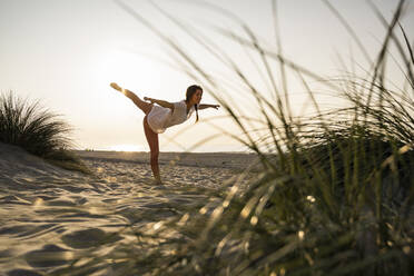 Flexible young woman practicing yoga on sand at beach during sunset - UUF21784